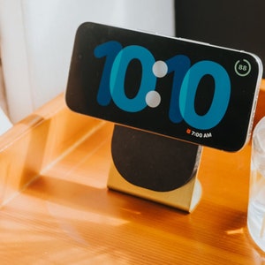 May include: A black smartphone with a blue and white digital clock face displaying 10:10. The phone is on a gold stand with a black base. The phone screen also displays "7:00 AM".