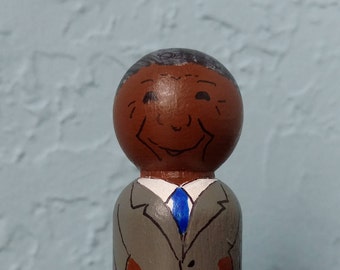 May include: A hand-painted wooden peg doll with a brown face, gray suit, and blue tie. The doll is smiling.