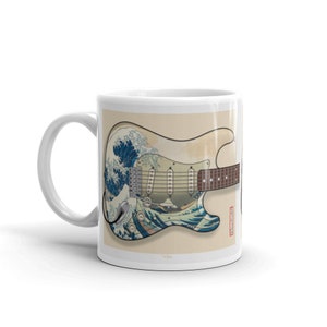 K&ouml;nnte beinhalten: Wei&szlig;e Keramiktasse mit einem Motiv einer Gitarre mit einem blauen und wei&szlig;en Wellendesign. Die Gitarre ist eine E-Gitarre im Stil einer Fender Stratocaster.