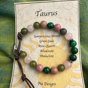 Puede incluir: Pulsera del zodíaco Tauro con cuentas de jade verde, cuarzo rosa, rodonita y malaquita. La pulsera está hecha con cordón de cuero marrón y un cierre plateado. La tarjeta dice "Tauro" y "Piedras semipreciosas: Jade verde, Cuarzo rosa, Rodonita, Malaquita" y "Pia Designs".