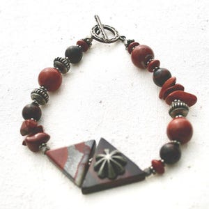 May include: A handmade bracelet with red, brown, and silver beads. The design incorporates round and irregular beads, with two triangular focal pieces. The clasp is a silver toggle.