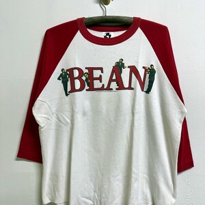 Op de afbeelding: Een wit baseball-stijl T-shirt met rode driekwart raglanmouwen en een rode ronde hals. De voorkant toont het woord BEAN in grote rode letters, met kleine afbeeldingen van een man in een pak geïntegreerd in het tekstontwerp.