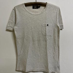 May include: A short-sleeved, light beige textured t-shirt with a crew neckline and a single chest pocket featuring a small dark button. The interior neck label displays the text RRL. The fabric has a subtle, uniform woven pattern throughout.