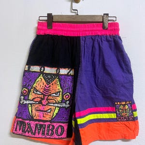 Op de afbeelding: Vintage sportbroek met een levendig kleurenschema. De broek heeft een zwart, paars en oranje ontwerp met een neonroze tailleband. Het linkerbeen heeft een afbeelding van een gezicht met het woord "MAMBO".