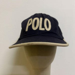 May include: Navy blue visor with the word "POLO" embroidered in a cream color across the front. The visor has a cream-colored trim along the brim.