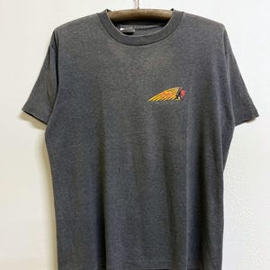 May include: A charcoal gray short-sleeve t-shirt featuring a small graphic on the left chest. The graphic depicts a yellow feathered headdress with the red text Indian and the word MOTOCYCLE printed in black underneath the design.