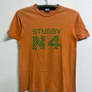 May include: An orange short-sleeve t-shirt featuring a green graphic print on the chest. The graphic displays the text STUSSY N°4, with the letters and numbers filled with a repeating pattern of small geometric shapes and symbols.