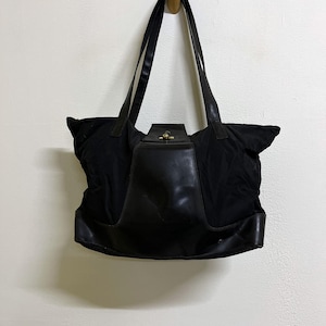May include: A black tote bag featuring a smooth, structured central panel and a matching base. The bag includes two long shoulder straps and a top flap closure secured with a small, circular gold-toned metal turn-lock fastener at the center.