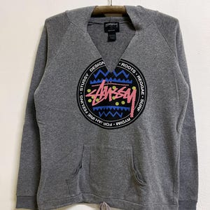 May include: Grey hoodie with a large circular graphic. The graphic features the word "Stussy" in a stylized font, surrounded by text and colourful geometric patterns. The hoodie has a drawstring at the waist and a front pocket.