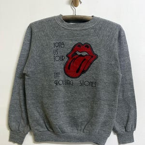 Può includere: Una felpa girocollo grigio melange con un logo a lingua dei Rolling Stones rosso e nero sul petto. Il testo recita 1978 US TOUR THE ROLLING STONES in caratteri neri. Il capo presenta maniche lunghe con polsini e orlo a costine.