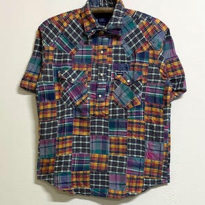 Puede incluir: Una camisa de manga corta con botones y un patrón de cuadros tipo patchwork multicolor en tonos amarillo, morado, verde azulado y negro. La camisa incluye un cuello puntiagudo, una tapeta frontal parcial con botones y dos bolsillos en el pecho.