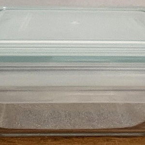 May include: A rectangular clear glass food storage container with a light blue plastic lid. The lid features a secure locking mechanism with four side flaps that fold down to seal the container, ensuring the contents remain contained and protected.
