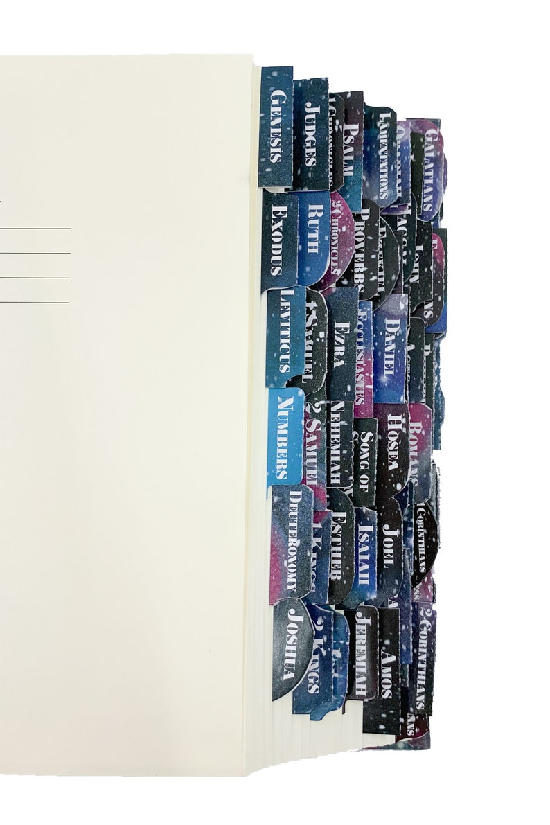 Op de afbeelding: Een boek met gekleurde tabbladen met de namen van bijbelboeken. De tabbladen hebben een galaxy-achtig ontwerp met witte tekst. De pagina's van het boek zijn wit en de tabbladen zijn in verschillende tinten blauw, paars en zwart.