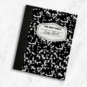 May include: A black and white composition notebook with the text "THE HOLY BIBLE" and the name "Emily Goode" in a cream-colored label. The notebook has a black spine and a speckled pattern.