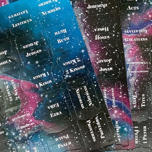 Op de afbeelding: Bijbeltabs met een galaxy-ontwerp. De tabs zijn bedrukt met de namen van de boeken van de Bijbel in witte letters. De achtergrond is een mix van blauw, paars en zwart met witte sterachtige spikkels.