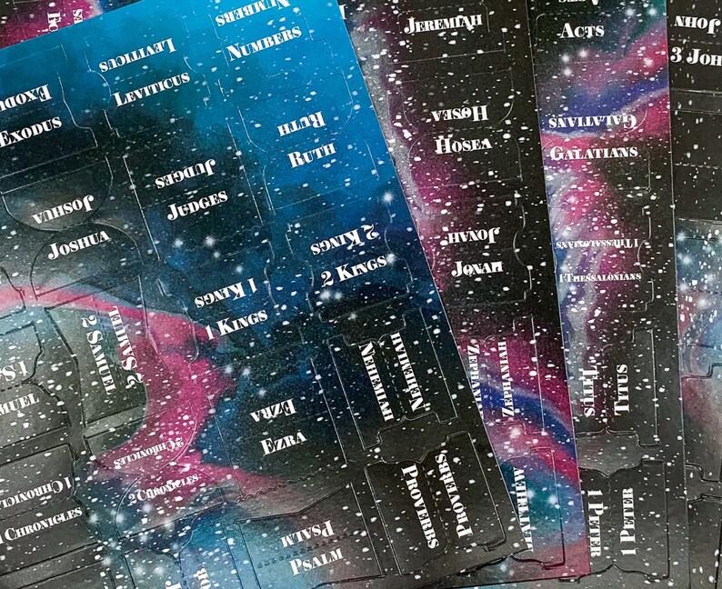 Op de afbeelding: Bijbeltabs met een galaxy-ontwerp. De tabs zijn bedrukt met de namen van de boeken van de Bijbel in witte letters. De achtergrond is een mix van blauw, paars en zwart met witte sterachtige spikkels.