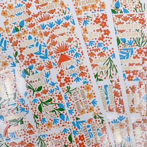 Op de afbeelding: Een verzameling Bijbel-tabs met bloemmotieven. De tabs hebben oranje, blauwe en groene bloempatronen met gouden letters. De tabs zijn gelabeld met verschillende namen van boeken uit de Bijbel, zoals "Rechters" en "Spreuken".