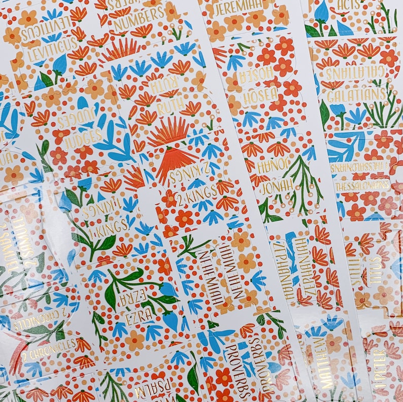 Op de afbeelding: Een verzameling Bijbel-tabs met bloemmotieven. De tabs hebben oranje, blauwe en groene bloempatronen met gouden letters. De tabs zijn gelabeld met verschillende namen van boeken uit de Bijbel, zoals "Rechters" en "Spreuken".