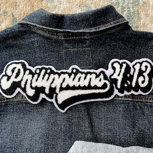 May include: A black and white embroidered patch with the text "Philippians 4:13" in a cursive font. The patch is sewn onto a blue denim jacket.
