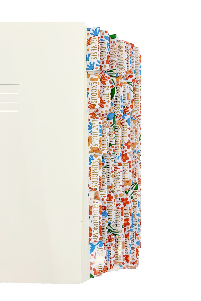 Op de afbeelding: Een cr&egrave;mekleurig boek met bloemmotief tabs. De tabs zijn gelabeld met gouden tekst, waaronder "Genesis", "Exodus" en "Leviticus". Het bloemmotief heeft rode, blauwe en oranje bloemen.