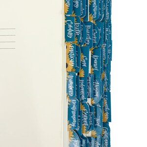 Op de afbeelding: Een boek met tealkleurige tabbladen met gouden zonnebloemen en witte letters. De tabbladen zijn gelabeld met boeknamen zoals Genesis, Exodus en Rechters. De pagina's van het boek zijn cr&egrave;mekleurig.