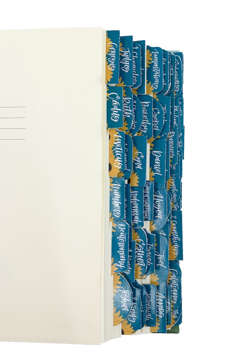 Op de afbeelding: Een boek met tealkleurige tabbladen met gouden zonnebloemen en witte letters. De tabbladen zijn gelabeld met boeknamen zoals Genesis, Exodus en Rechters. De pagina's van het boek zijn cr&egrave;mekleurig.