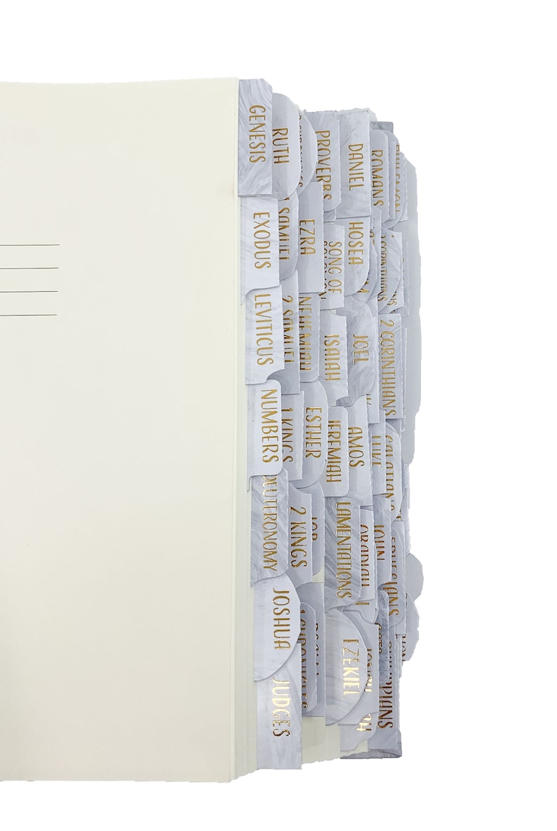 Op de afbeelding: Cr&egrave;mekleurige ordner met meerdere gelabelde tabbladen. De tabbladen zijn wit met een marmerontwerp en gouden letters, met verschillende boektitels uit de Bijbel. Het bestand is verticaal geplaatst.