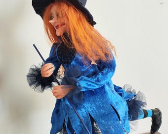 Könnte beinhalten: Eine blaue und schwarze Hexenpuppe mit roten Haaren und einem schwarzen spitzen Hut. Die Puppe hält einen schwarzen Besen und steht auf einem schwarzen Sockel.