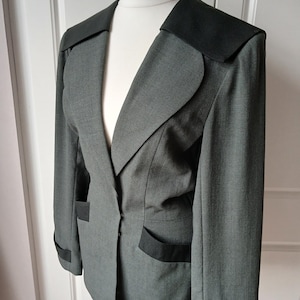 May include: A charcoal grey blazer featuring a wide black collar, black pocket flaps, and black cuffs on the long sleeves. The jacket has a structured silhouette with a single-button closure at the waist and a smooth, solid-coloured fabric finish.