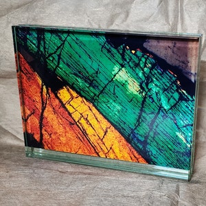 May include: A rectangular glass block displaying an abstract design. The artwork is split diagonally, with orange and yellow on one side and green and blue on the other, reminiscent of a geological formation.