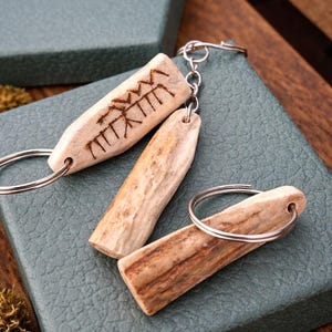 Könnte beinhalten: Drei Schlüsselanhänger aus naturfarbenem Holz, jeweils mit einem silberfarbenen Ring. Ein Schlüsselanhänger hat ein geschnitztes Design. Die Schlüsselanhänger werden auf einer strukturierten, teal-farbenen Oberfläche mit einer teal-farbenen Box im Hintergrund präsentiert. Der Text "LindenHand Crafts" ist sichtbar.