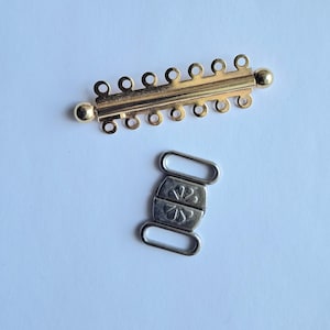 Peut inclure: Deux fermoirs de bijoux. L'un est doré, rectangulaire, avec de multiples petites boucles et des extrémités arrondies. L'autre est argenté, avec un motif floral décoratif et deux boucles rectangulaires. Les deux fermoirs sont sur fond blanc.