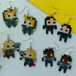 May include: A collection of pixelated character earrings with silver hooks. The earrings feature various characters in shades of blue, yellow, brown, and red. The earrings are displayed on a white surface with yellow and orange flowers.