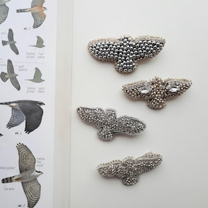 May include: Four handmade bird-shaped brooches. The brooches are embellished with silver beads and crystals. One brooch has two clear crystals. The brooches are displayed on a white surface next to a book with bird illustrations.