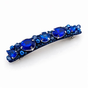 May include: A blue hair barrette adorned with large, oval and round blue gemstones. Smaller blue beads and teardrop-shaped gems surround the larger stones. The barrette is set on a dark blue base with a silver clasp.