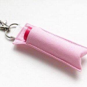 May include: A light pink fabric lip balm holder with a silver-colored metal clip. The holder has a white strap and a pink lip balm is partially visible. The holder is designed to protect and carry lip balm.