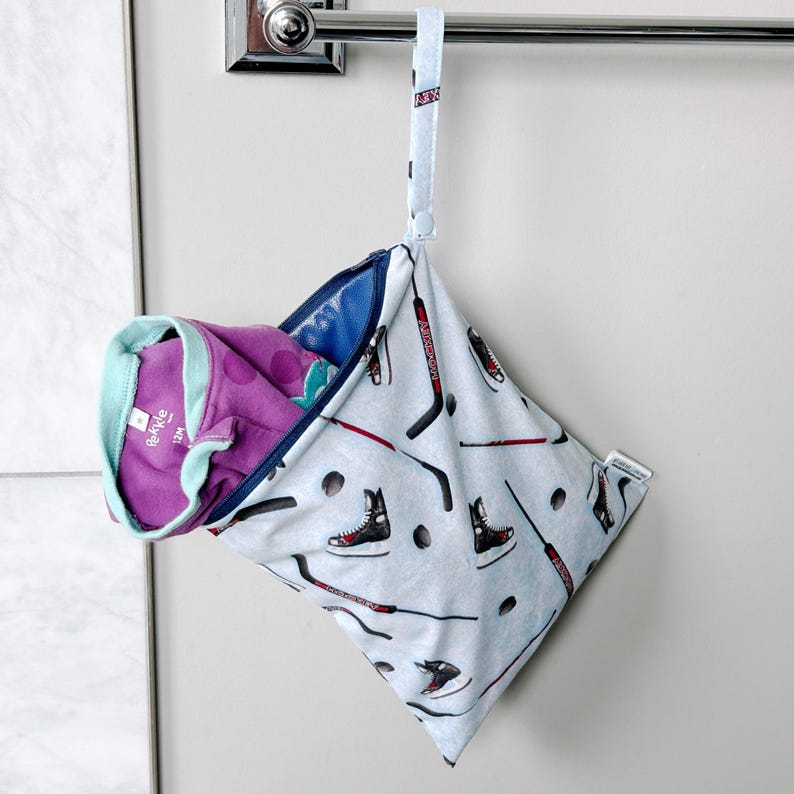 May include: A light blue, hockey-themed wet bag with a zipper, hanging from a chrome towel bar. The bag features a pattern of hockey sticks, skates, and pucks. Inside, a purple and teal garment is visible.