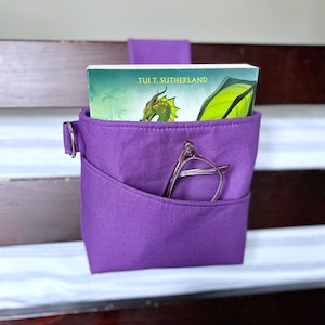 May include: A purple fabric bedside caddy featuring a front pocket containing a pair of glasses. A book titled Tui T. Sutherland with a green dragon illustration is tucked inside the main compartment. The caddy has a metal ring on the side.