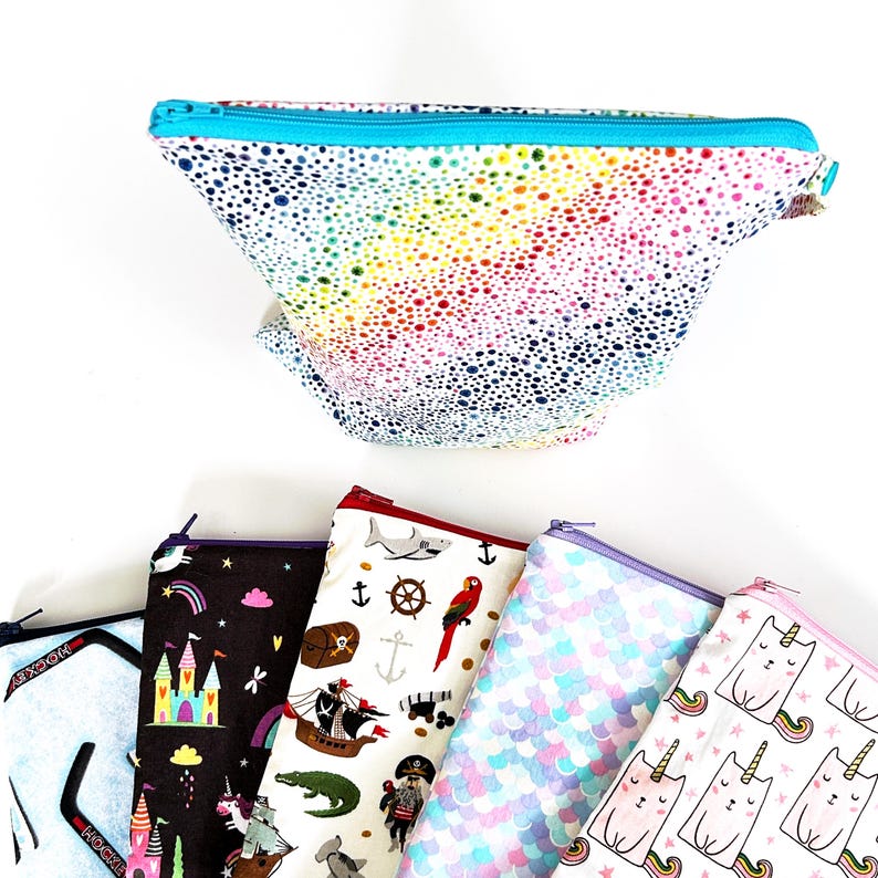 May include: A collection of zippered pouches with various designs. One pouch features a rainbow dotted pattern with a turquoise zipper. Other pouches display patterns of unicorns, pirates, mermaids, and cats with rainbows.