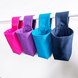 May include: Four colorful fabric hanging organizers in shades of pink, purple, turquoise, and navy blue. Each has a loop for hanging and a cylindrical pocket. The organizers are suspended from a white rod.