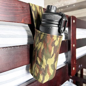 May include: A black water bottle with a black clip is held in a camouflage fabric holder. The holder is attached to a dark brown wooden bunk bed frame. The camouflage pattern includes shades of green, brown, and tan.