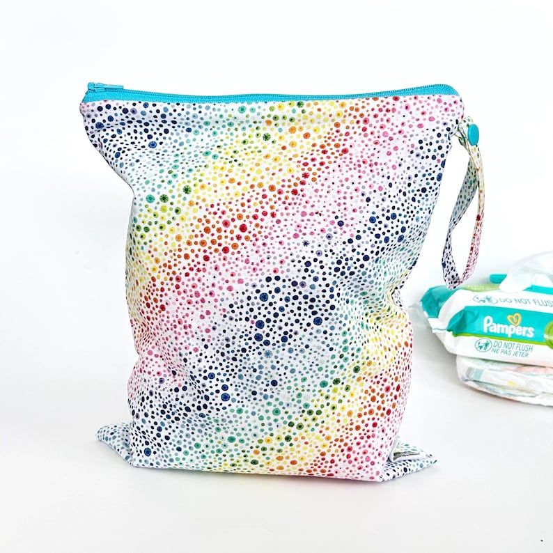 May include: A reusable, waterproof diaper bag with a colorful polka dot pattern in rainbow hues. The bag has a turquoise zipper and a matching handle. A stack of diapers is visible in the background.