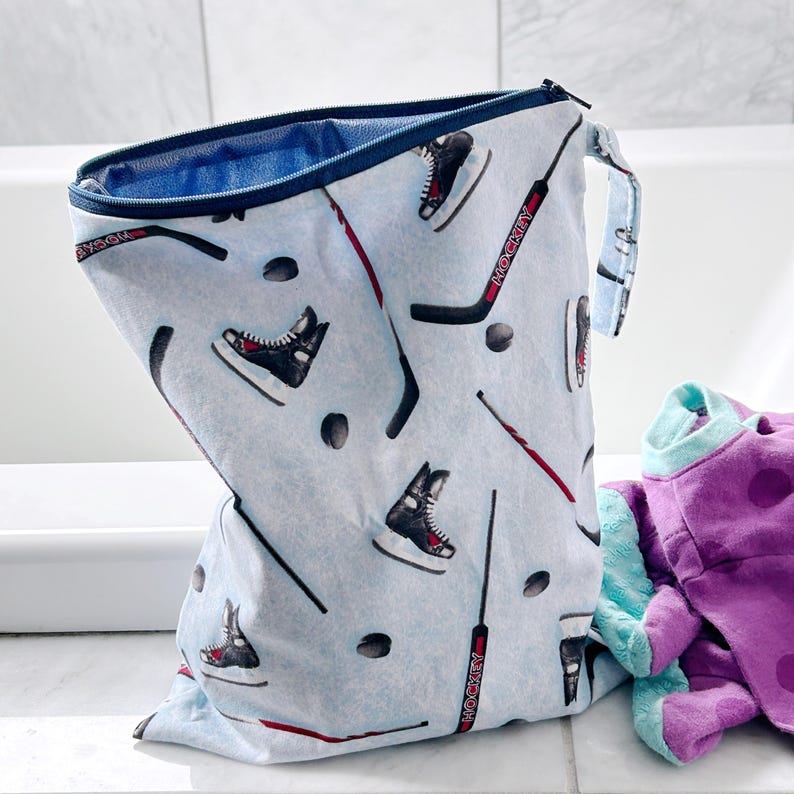 May include: A light blue, zippered bag with a hockey theme. The bag features a pattern of black hockey skates, pucks, and hockey sticks with the word "HOCKEY" printed on them. The interior is blue, and a purple and teal item is next to the bag.