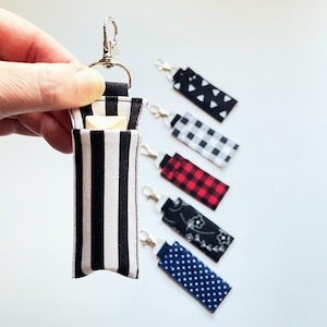 May include: A fabric keychain with a black and white striped pattern, holding a small yellow envelope. Several other keychains with various patterns, including black and white triangles, gingham, red plaid, floral, and blue polka dots, are also visible.