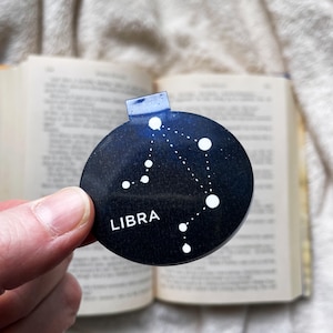 Könnte beinhalten: Runder, dunkelblauer Lesezeichen mit dem Sternzeichen Waage und dem Wort "LIBRA" in Weiß. Das Lesezeichen wird vor einem offenen Buch gehalten.