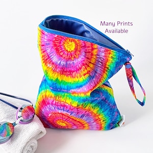 May include: A zippered pouch featuring a vibrant rainbow tie-dye pattern with circular motifs, a blue interior lining, and a matching wrist strap. The text Many Prints Available is printed in purple. Swimming goggles and a white towel are nearby.