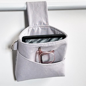 May include: A light grey fabric hanging storage organiser with a top strap loop. The main compartment holds a black e-reader, while a smaller front pocket contains a pair of pink-framed spectacles. A silver metal D-ring is attached to the side.