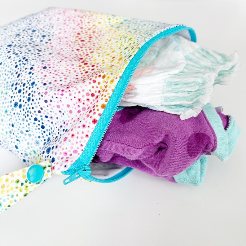 May include: A colorful, polka-dotted wet bag with a turquoise zipper and handle. The bag is open, revealing diapers and purple and teal clothing. The bag's pattern includes a rainbow of colors.