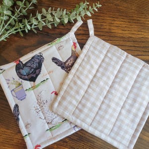 May include: Two quilted pot holders. One is white with a brown gingham pattern. The other is white with a chicken and floral print. The text on the chicken print pot holder reads "White Frizzle" and "Australorp".
