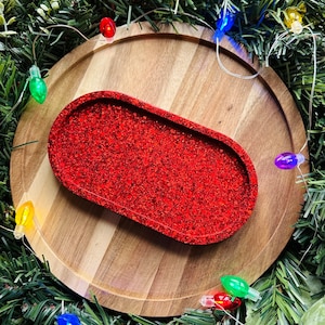 May include: A red glitter tray in an oval shape sits on a wooden round tray, surrounded by Christmas garland and string lights. The tray is a vibrant, eye-catching accent piece.